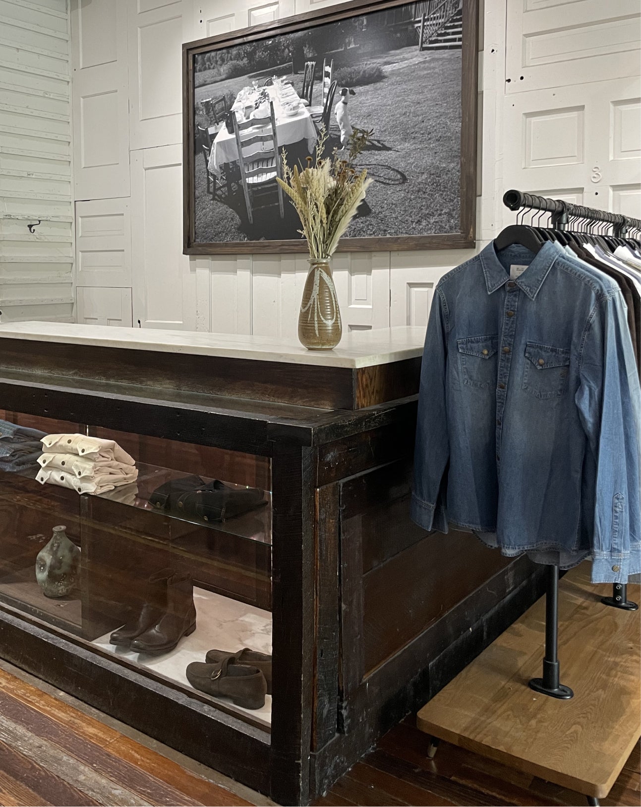 A blue denim button-up shirt hangs on a clothing rack next to a dark wooden display counter in a boutique setting. A framed black-and-white photograph and a vase with dried grass are on the counter, with shoes displayed inside the glass-front lower cabinet.