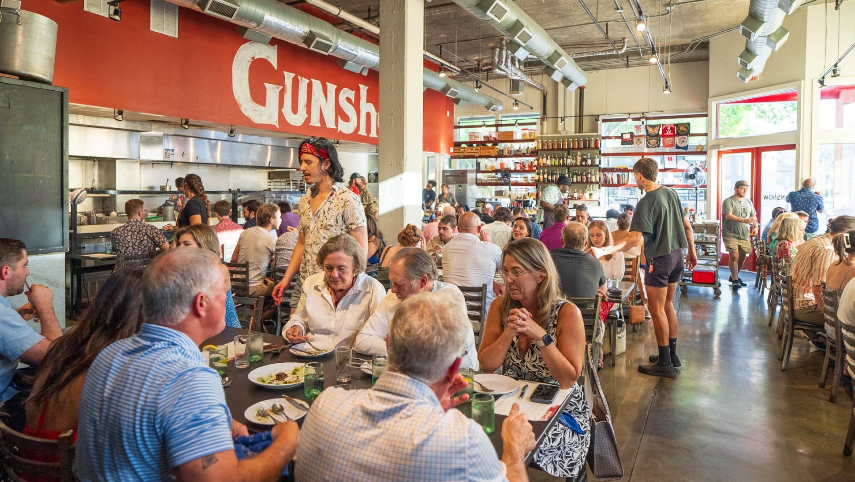 A busy restaurant dining area with multiple people seated at tables and standing in a kitchen/open kitchen space. A large red wall sign with white text reading 'Gunsh' is visible above the seating area.