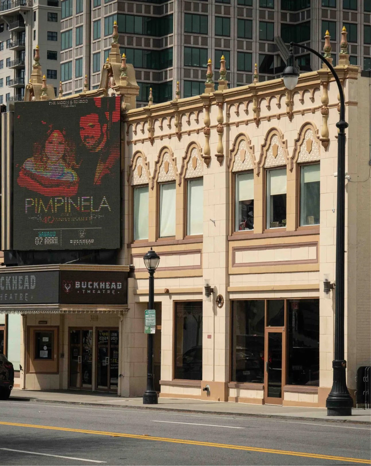 A city street scene showing a beige ornate building facade with storefront windows, a large dark movie poster reading PIMPINELA on the left, and a Buckhead Theatre sign below. Tall modern buildings are visible in the background behind the structure.