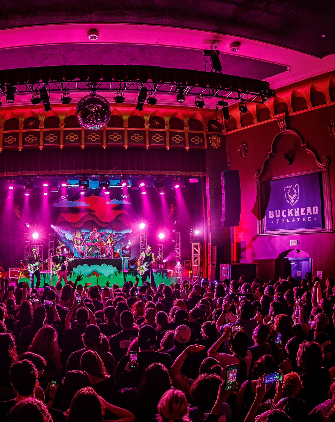 A live concert performance at Buckhead Theatre, with a band on stage under pink and purple stage lights, a large audience in the foreground, and a screen displaying the Buckhead Theatre logo.