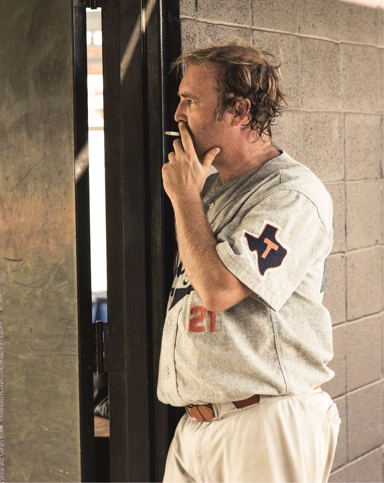A man in a distressed gray baseball jersey with a Texas 'T' patch on the sleeve and the number 21 on the front, leaning against a cinderblock wall and smoking a cigarette.