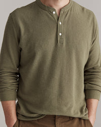 A person wearing an olive green, long-sleeve Henley shirt with three white buttons, and brown pants. The persons face is partially out of frame.