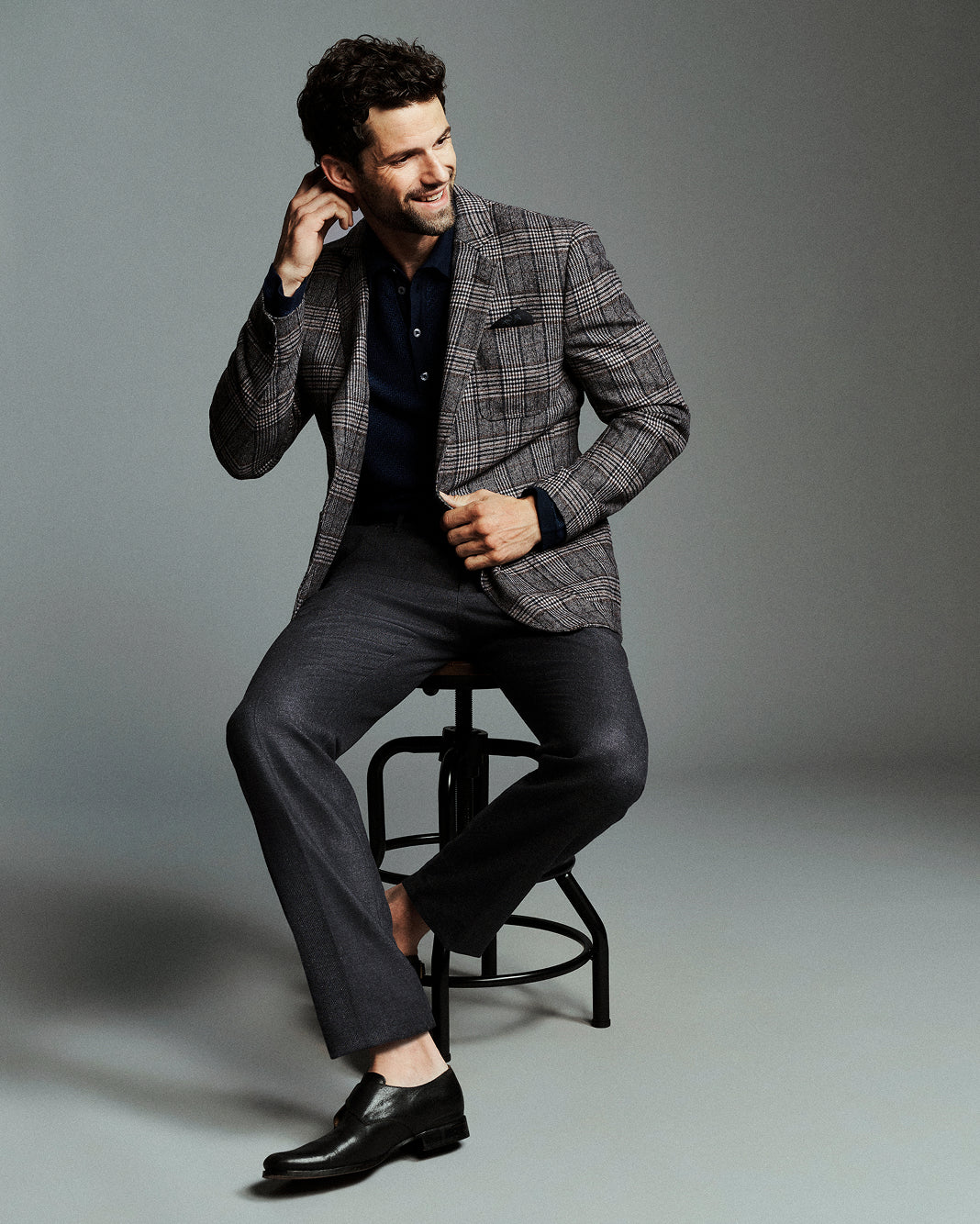 A man wearing a gray and black plaid blazer, dark trousers, and black dress shoes sits on a black stool against a gray studio background.