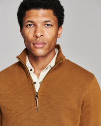 A man wearing a brown quarter-zip pullover sweater over a cream button-down shirt, photographed against a solid light gray background.