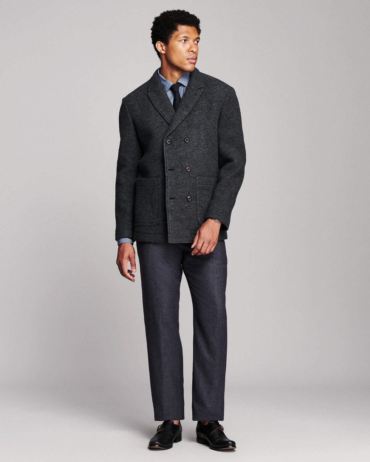 A man modeling a dark gray double-breasted wool-blend blazer with matching gray trousers, a light blue dress shirt, a dark tie, and black loafers, photographed against a neutral light gray studio background.