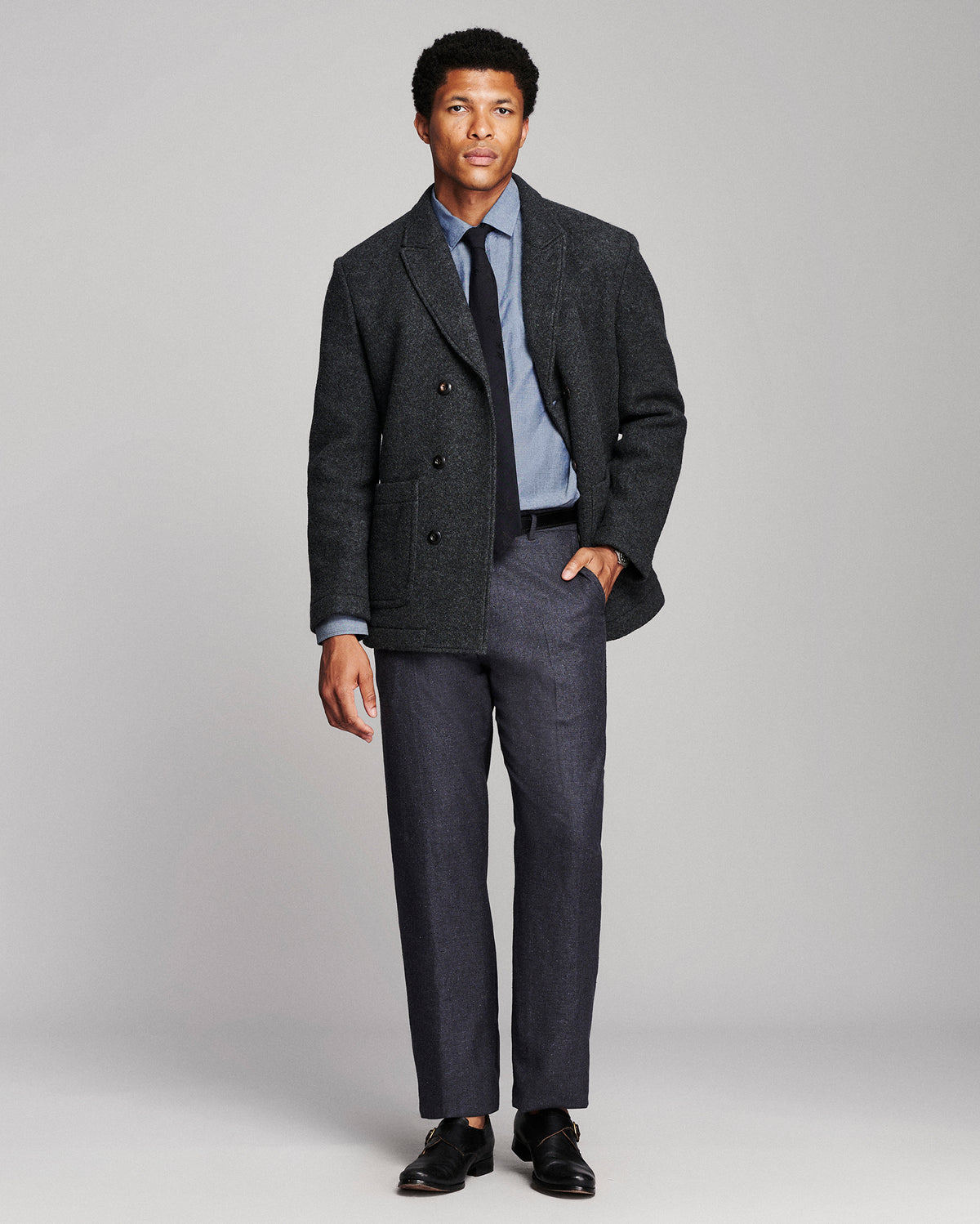 A man modeling a dark charcoal gray double-breasted blazer with matching gray trousers, a light blue dress shirt, a dark tie, and black dress shoes, photographed against a neutral light gray studio background.