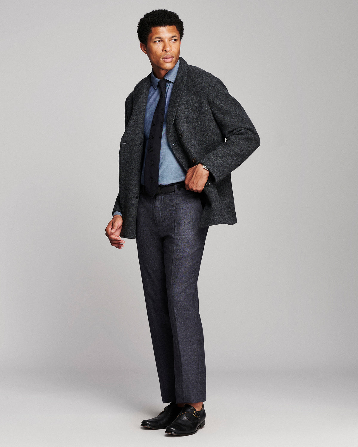 A man modeling a dark charcoal tweed blazer, dark gray trousers, a light blue dress shirt, a navy tie, and black dress shoes against a light gray studio background.