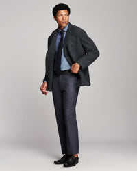 A man modeling a dark charcoal tweed blazer, dark gray trousers, a light blue dress shirt, a navy tie, and black dress shoes against a light gray studio background.