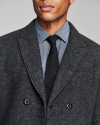 Close-up of a man wearing a dark charcoal gray double-breasted wool-blend overcoat, styled with a light blue chambray shirt and a black patterned tie, against a neutral gray background.