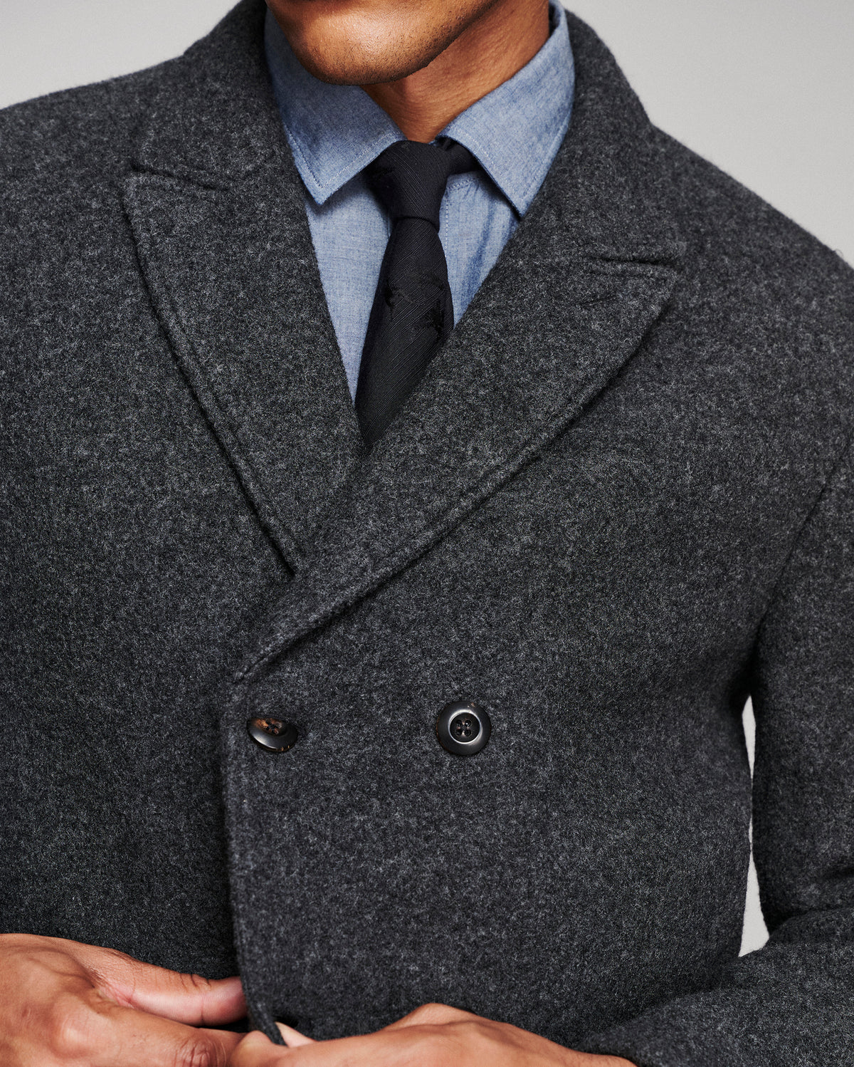 Close-up of a man wearing a dark charcoal gray double-breasted wool-blend overcoat, styled with a light blue dress shirt and a black tie.