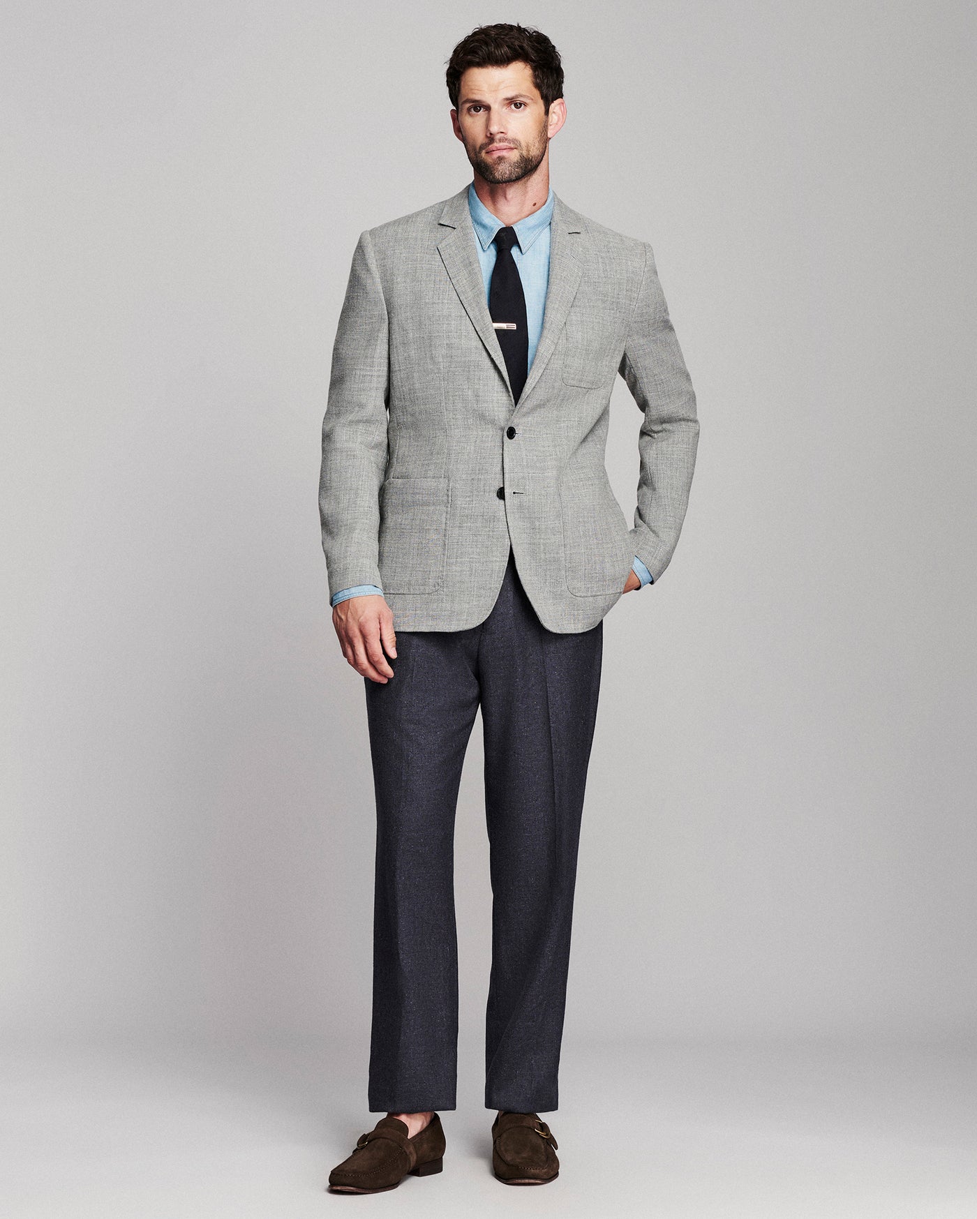 A man modeling a light gray textured blazer with a light blue dress shirt, black tie, dark gray trousers, and brown suede loafers, standing against a neutral gray studio background.