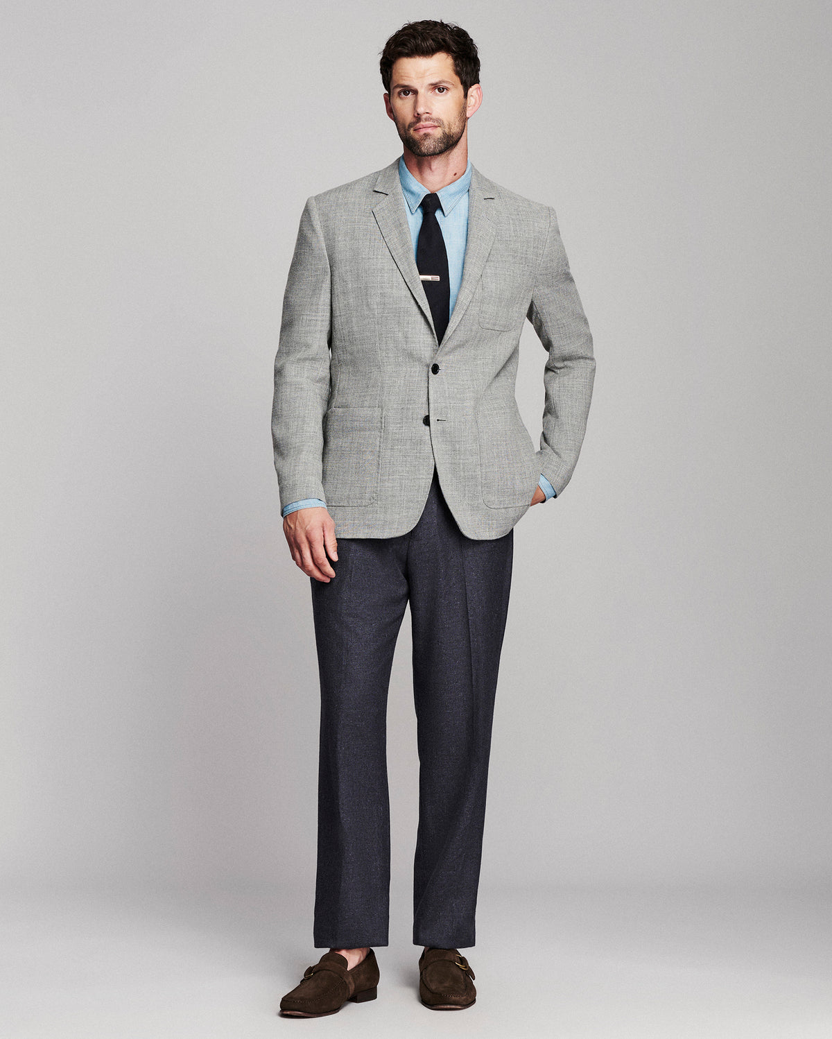 A man modeling a light gray textured blazer with a light blue dress shirt, black tie, dark gray trousers, and brown suede loafers, standing against a neutral gray studio background.