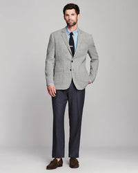 A man modeling a light gray textured blazer with a light blue dress shirt, black tie, dark gray trousers, and brown suede loafers, standing against a neutral gray studio background.