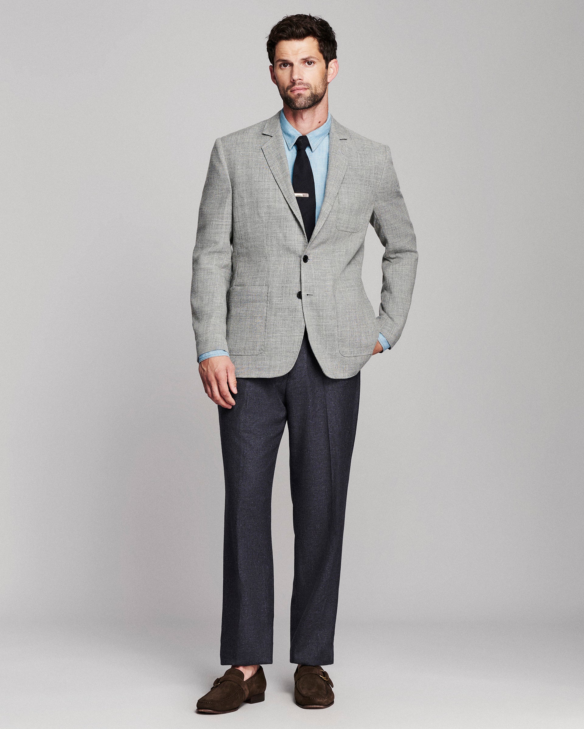 A man modeling a light gray textured blazer with a light blue dress shirt, black tie, dark gray trousers, and brown suede loafers, standing against a neutral gray studio background.