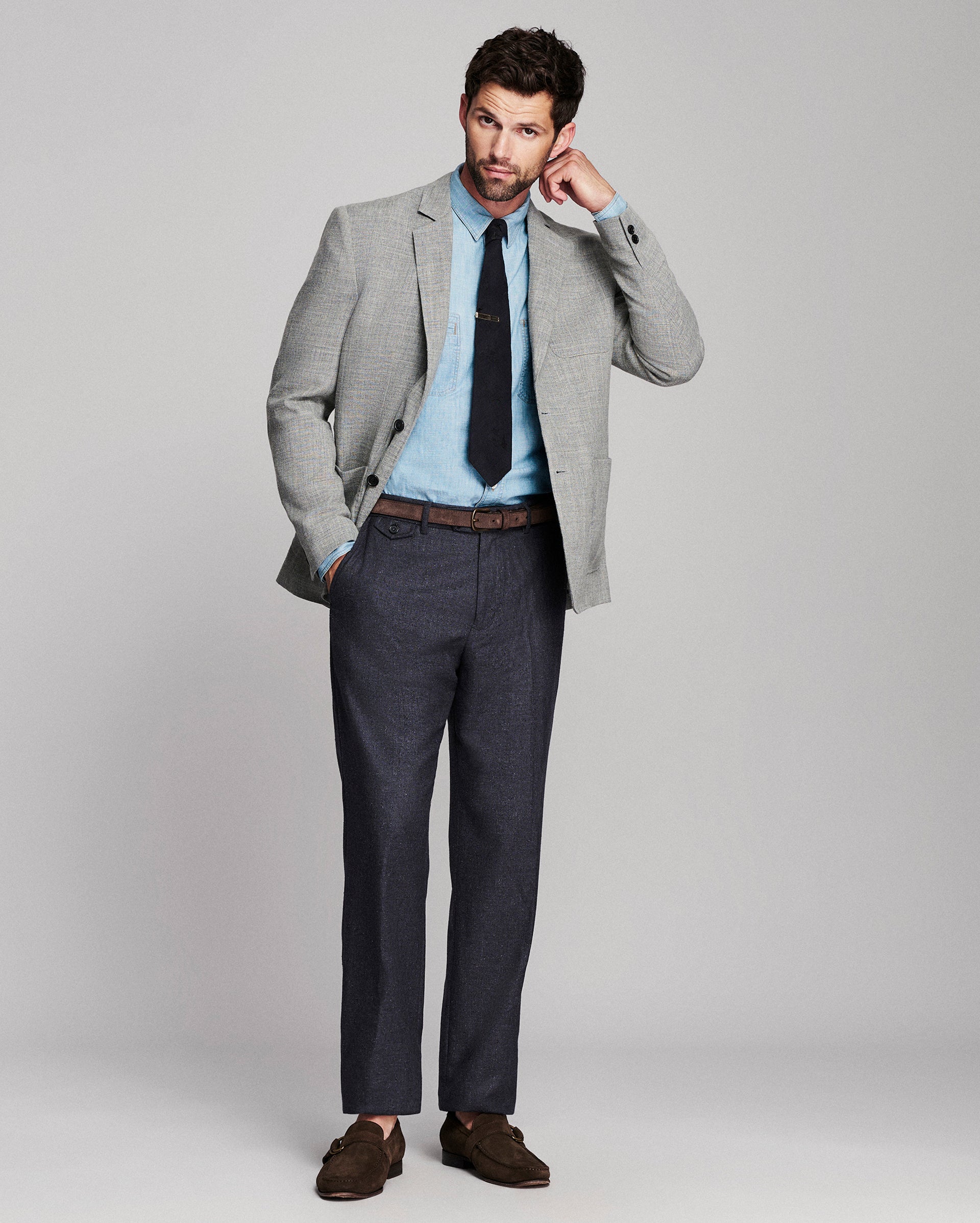 A man modeling a light gray blazer, light blue dress shirt, black tie, dark gray trousers, and brown loafers against a light gray studio background.