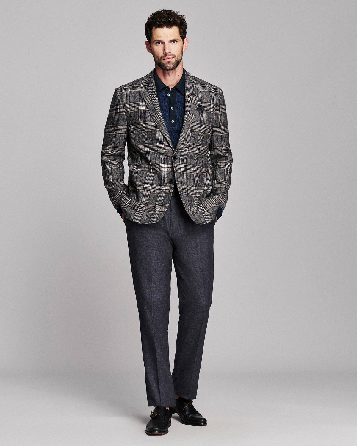 A man modeling a gray and navy plaid blazer with matching gray trousers, a navy polo shirt, and black dress shoes, standing against a light gray studio background.