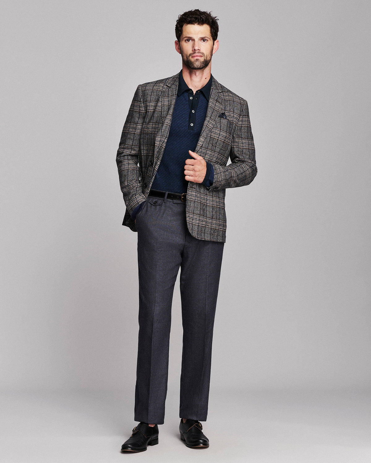 A man modeling a men's blazer with a gray and brown plaid pattern, worn over a dark navy polo shirt and paired with matching gray trousers, black dress shoes, and a black belt against a light gray studio background.