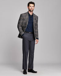 A man modeling a gray and black plaid blazer over a navy polo shirt, paired with charcoal gray trousers and black dress shoes, against a solid light gray studio background.