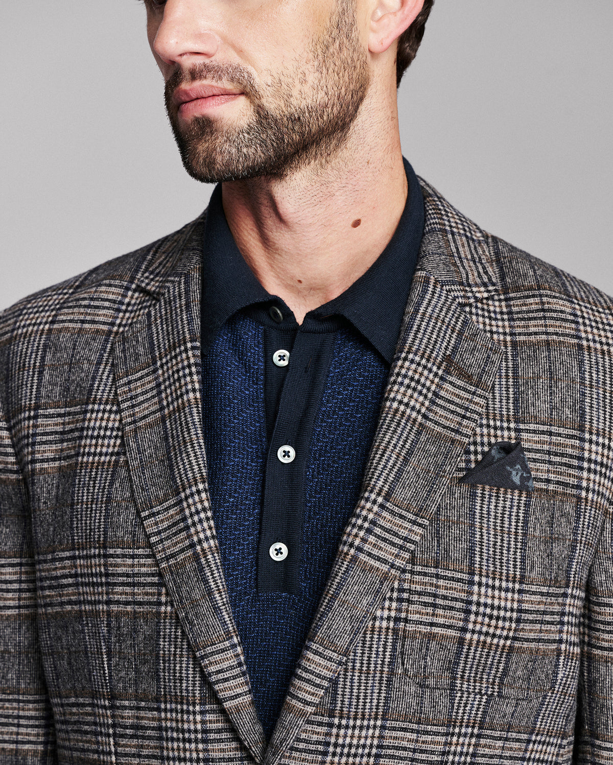 A close-up of a man wearing a gray and brown plaid blazer over a dark navy polo shirt with a matching pocket square.