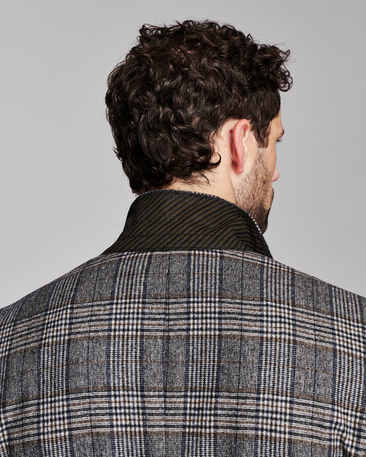 Back view of a man wearing a gray and black plaid wool blazer with a dark ribbed knit collar, set against a solid light gray background.