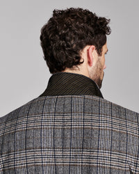 Back view of a man wearing a gray and black plaid wool blazer with a dark ribbed knit collar, set against a solid light gray background.