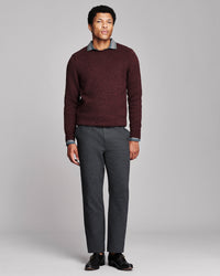 A man modeling a dark burgundy knit crewneck sweater with a gray collared shirt underneath, paired with dark gray trousers and black leather shoes, against a solid light gray studio background.