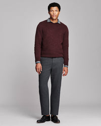 A man modeling a dark burgundy knit crewneck sweater layered over a gray collared shirt, paired with charcoal gray trousers and black dress shoes, against a solid light gray studio background.