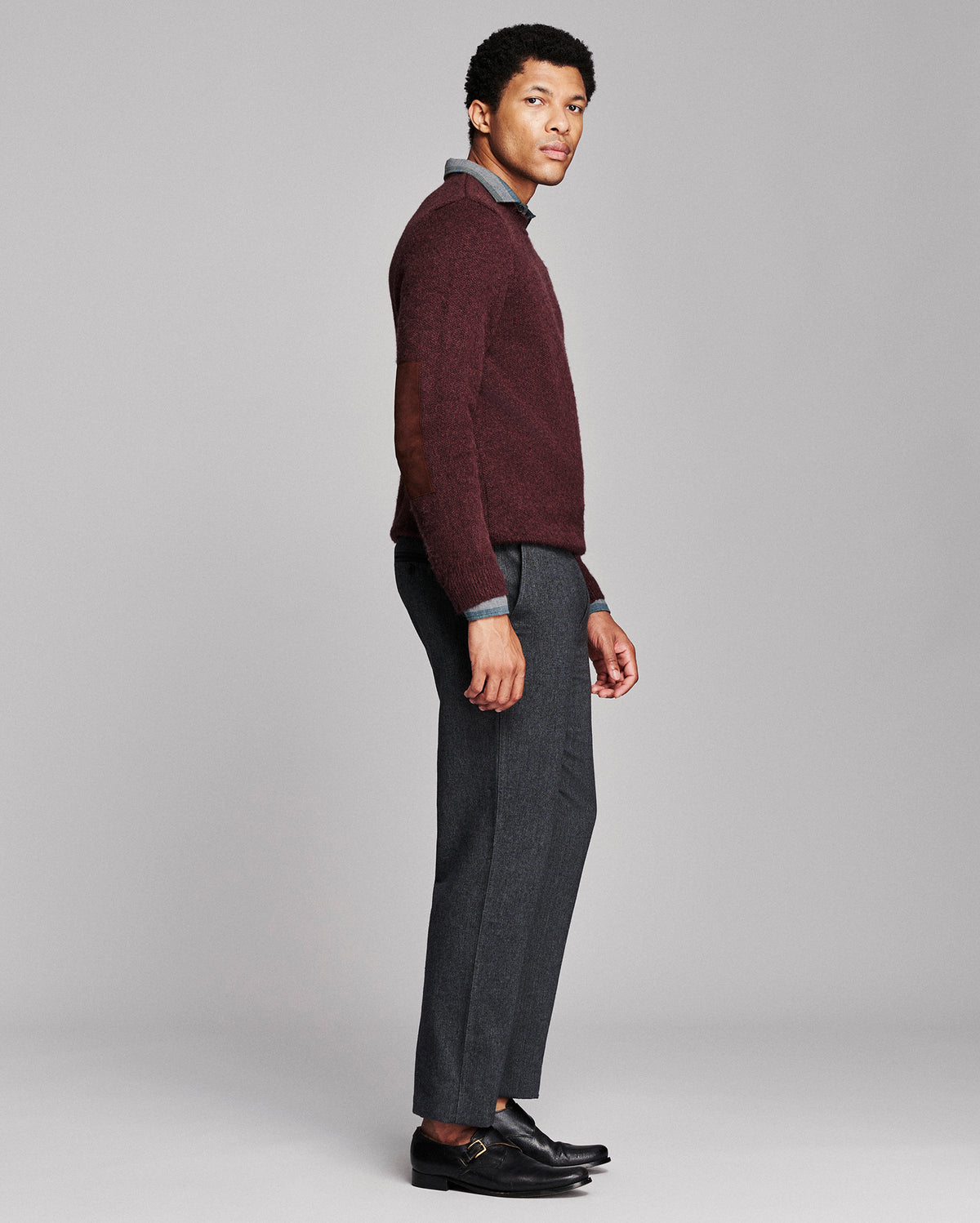 A man modeling a burgundy knit sweater with brown leather elbow patches, worn over a light blue collared shirt and paired with dark gray trousers and black loafers, against a solid gray studio background.