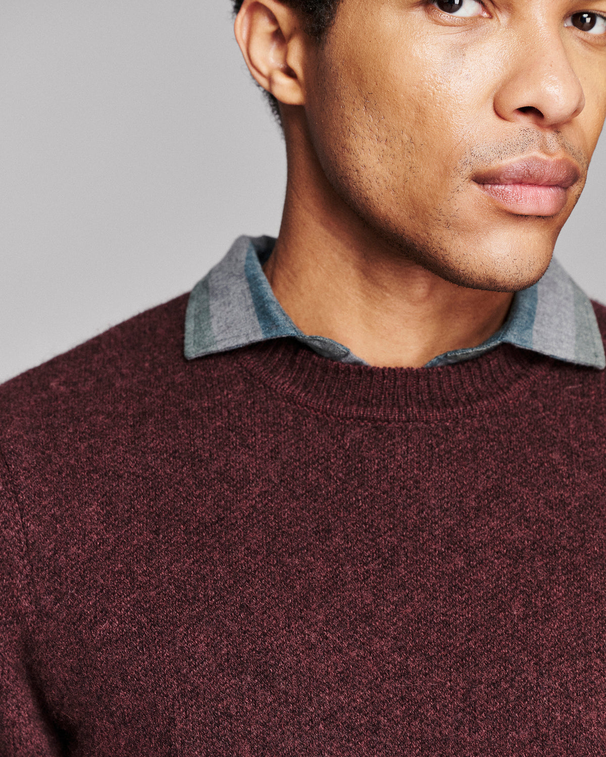 A close-up of a man wearing a textured burgundy knit crewneck sweater layered over a blue-gray collared shirt, against a neutral gray background.