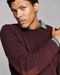 A close-up of a man wearing a maroon knit sweater with brown elbow patches and a light blue collared shirt underneath, set against a solid gray background.