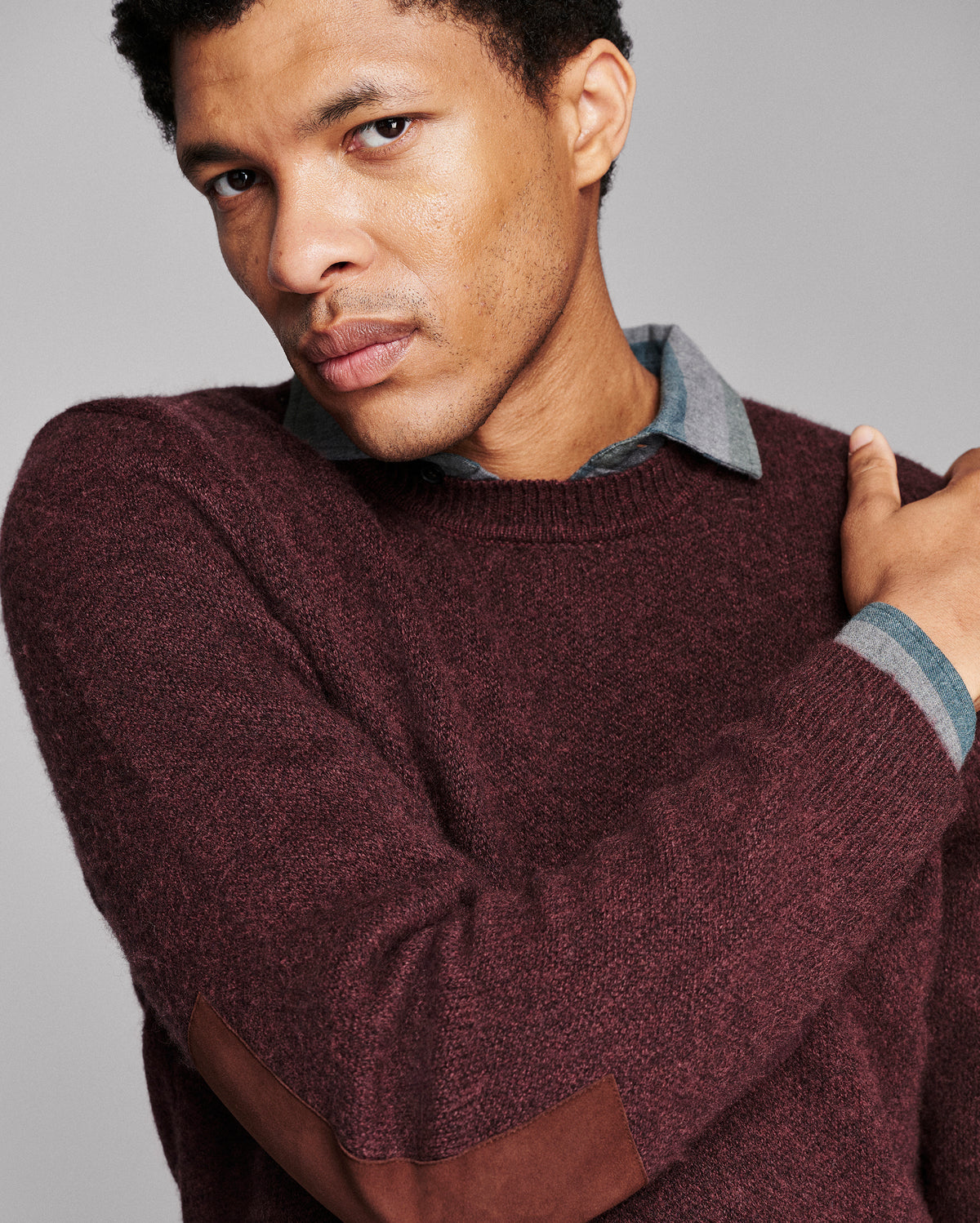 A close-up of a man wearing a maroon knit sweater with brown elbow patches and a light blue collared shirt underneath, set against a solid gray background.
