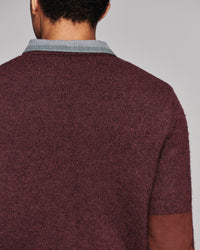 Close-up back view of a person wearing a short-sleeve knit sweater in heathered burgundy, with a light gray ribbed collar and brown suede-like elbow patches, against a plain light gray background.