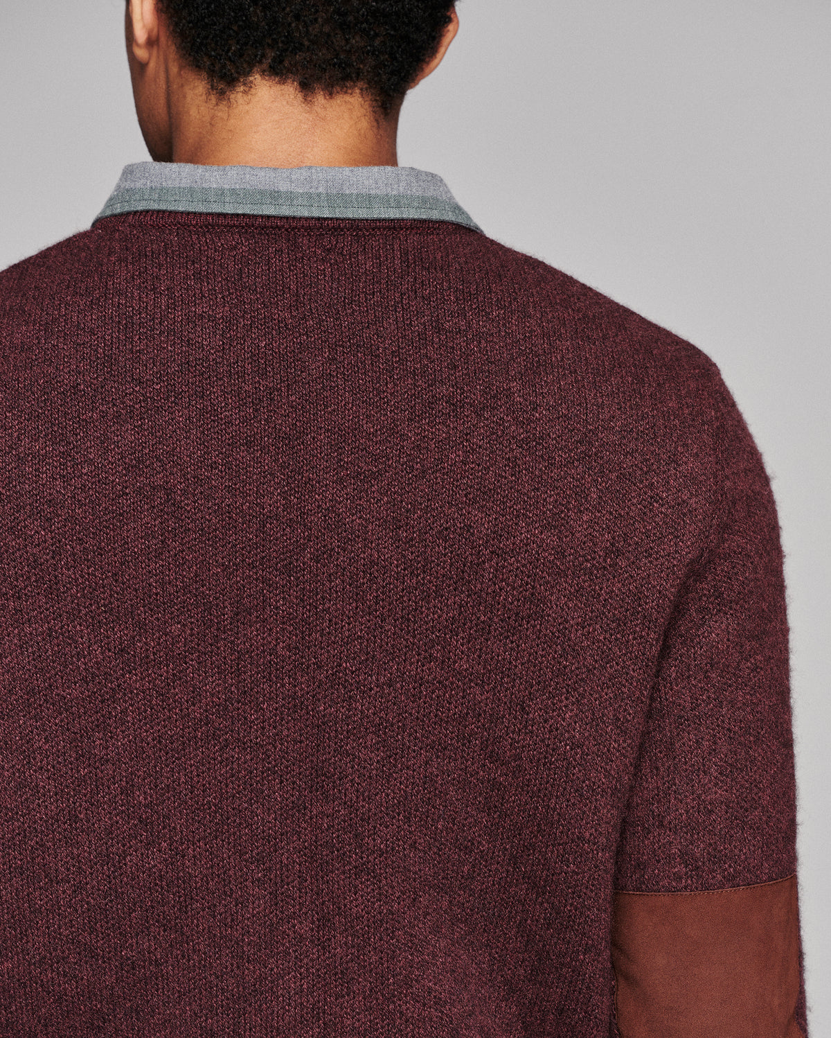 Close-up back view of a person wearing a short-sleeve knit sweater in heathered burgundy, with a light gray ribbed collar and brown suede-like elbow patches, against a plain light gray background.