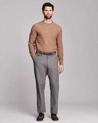 A man modeling a tan knit crewneck sweater with a subtle tonal fox pattern, paired with gray trousers, a brown belt, and brown shoes, against a solid light gray background.