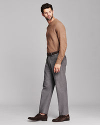 A man modeling a brown knit crewneck sweater with a subtle tonal pattern, paired with gray trousers and dark brown suede loafers, shot against a solid gray studio background.