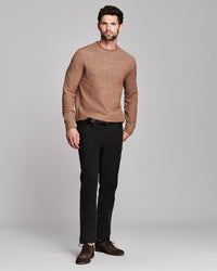 A man modeling a tan crewneck sweater with a subtle tonal pattern, paired with black trousers and brown leather shoes, against a solid light gray background.