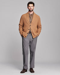 A man modeling a tan cable-knit shawl-collar cardigan over a patterned shirt and gray trousers, standing against a light gray studio background.