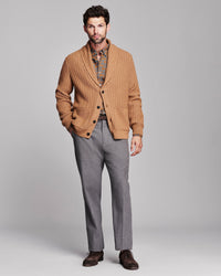 A man modeling a tan cable-knit shawl-collar cardigan over a patterned shirt, paired with gray trousers and brown leather shoes, against a solid light gray studio background.