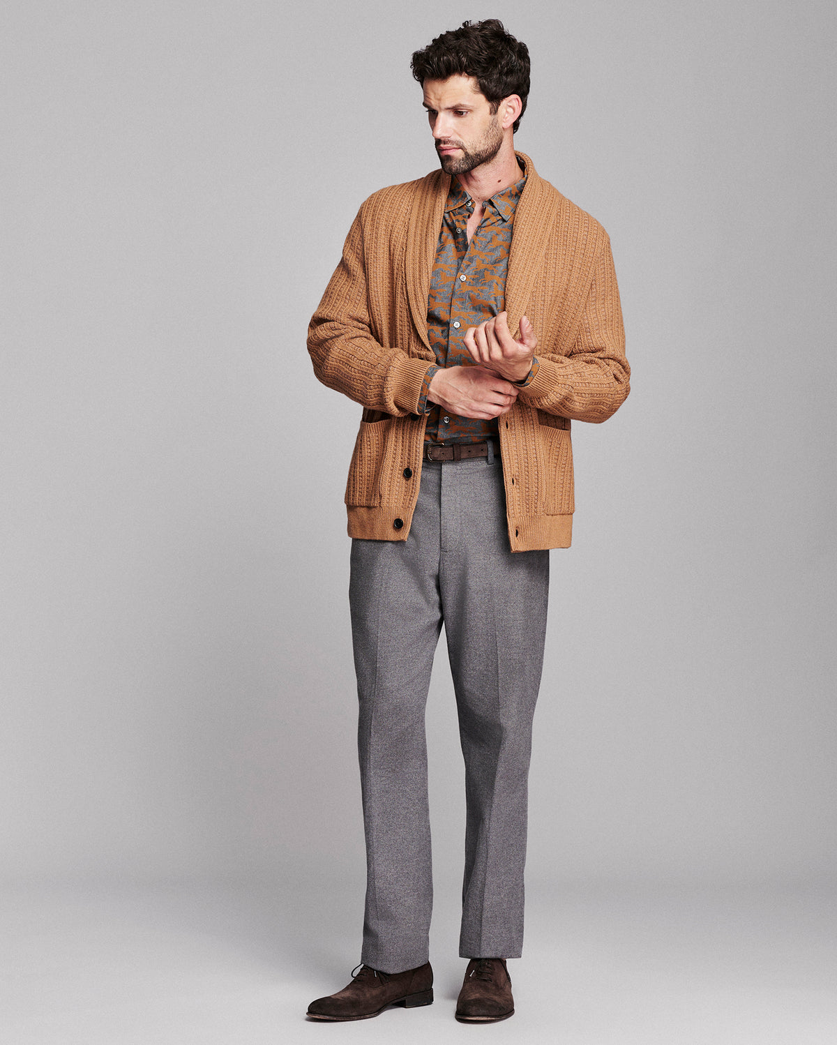 A man modeling a tan corduroy cardigan over a patterned shirt, paired with gray trousers and brown leather shoes, against a solid light gray studio background.