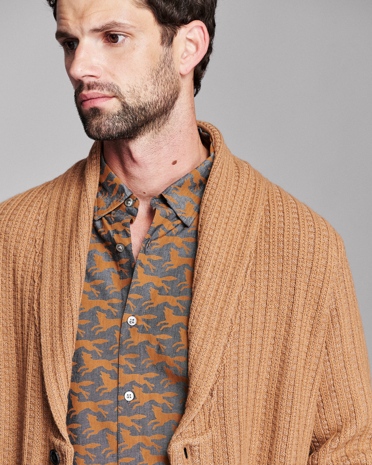 A close-up of a man wearing a tan ribbed knit cardigan over a brown and gray patterned button-down shirt, set against a solid gray studio background.