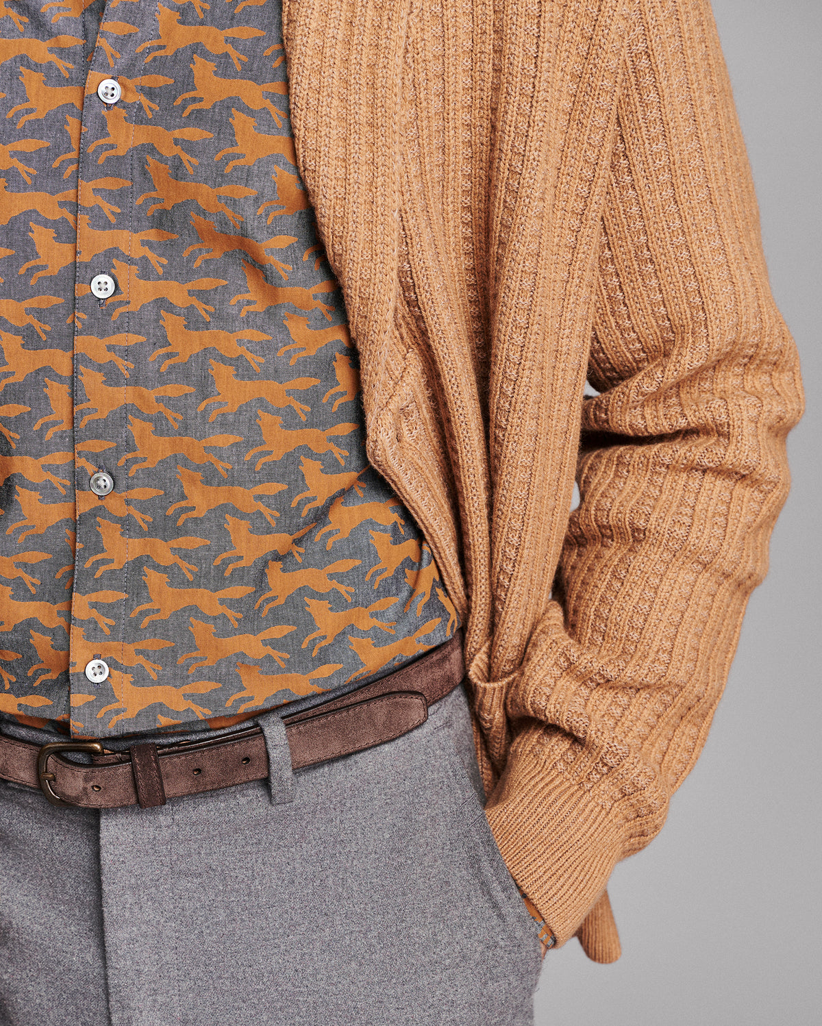 A close-up of a person wearing a tan ribbed knit cardigan over a brown and orange horse-patterned button-down shirt, paired with gray trousers and a brown belt, against a solid gray background.