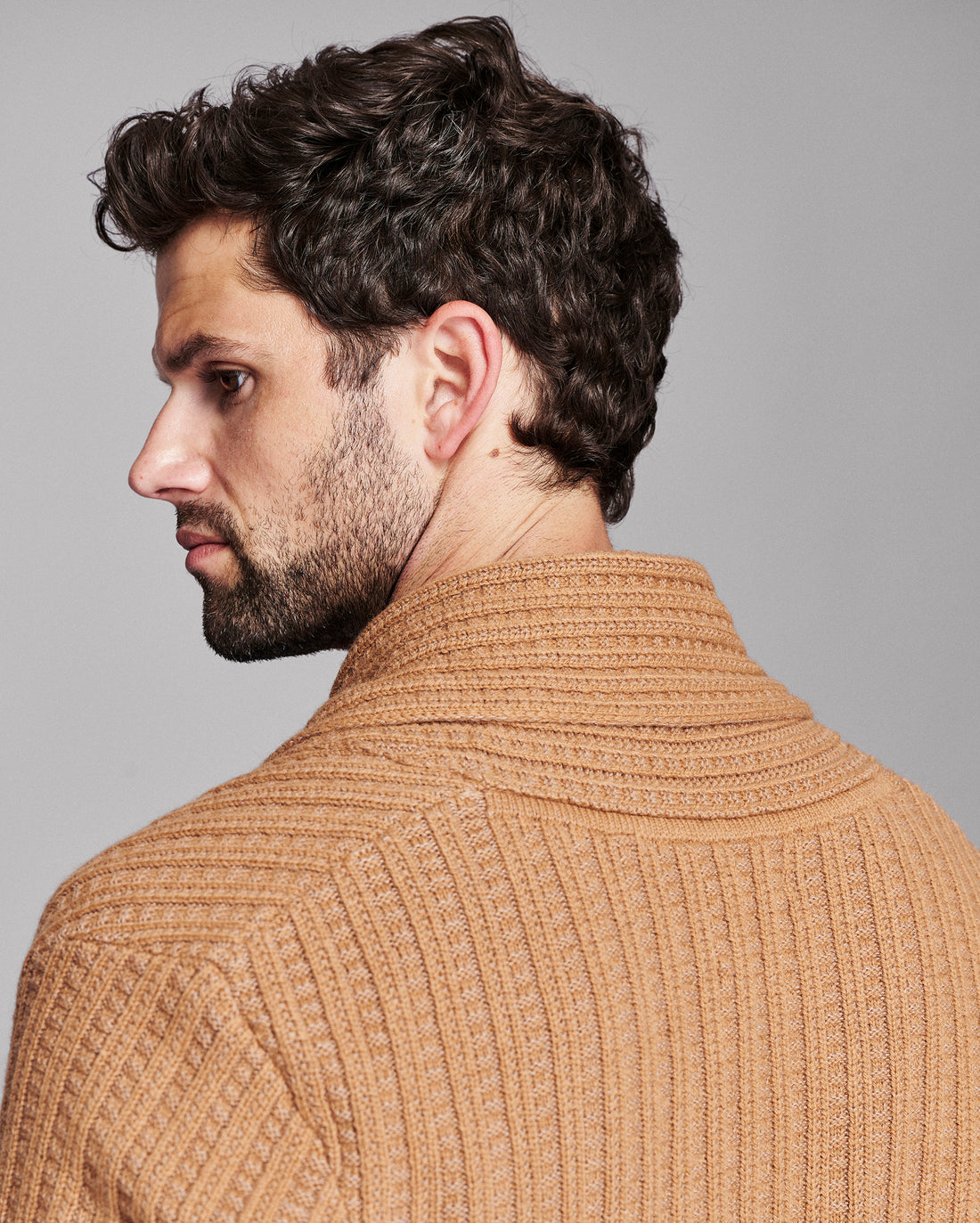 A man shown from the back and side profile wearing a ribbed knit sweater in a warm camel tan color against a solid gray studio background.