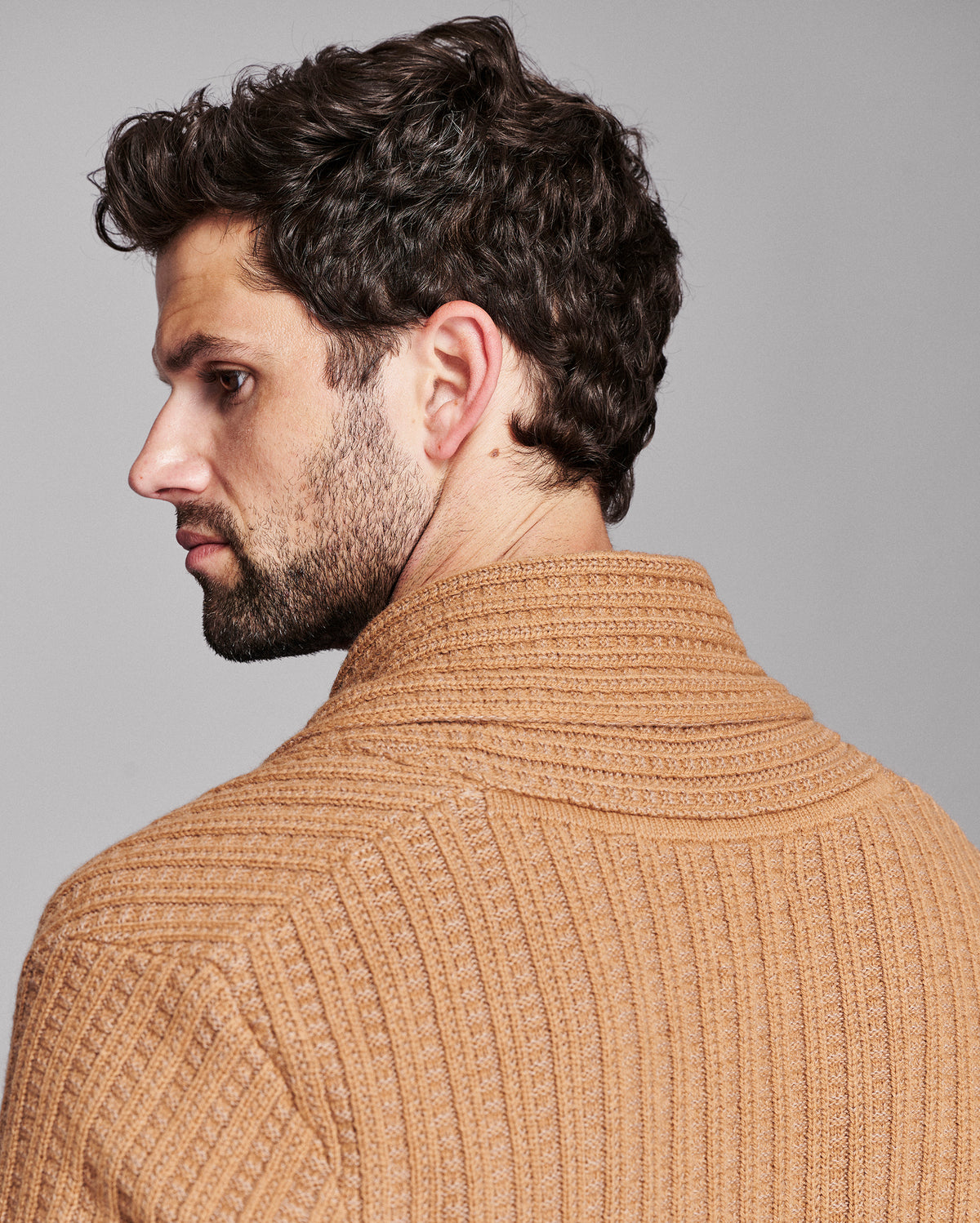 A man shown from the back and side profile wearing a ribbed knit sweater in a warm camel tan color against a solid gray studio background.
