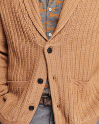 A close-up of a ribbed knit cardigan in warm camel brown, worn over a patterned shirt and gray trousers. The cardigan has black buttons and a shawl-style collar.
