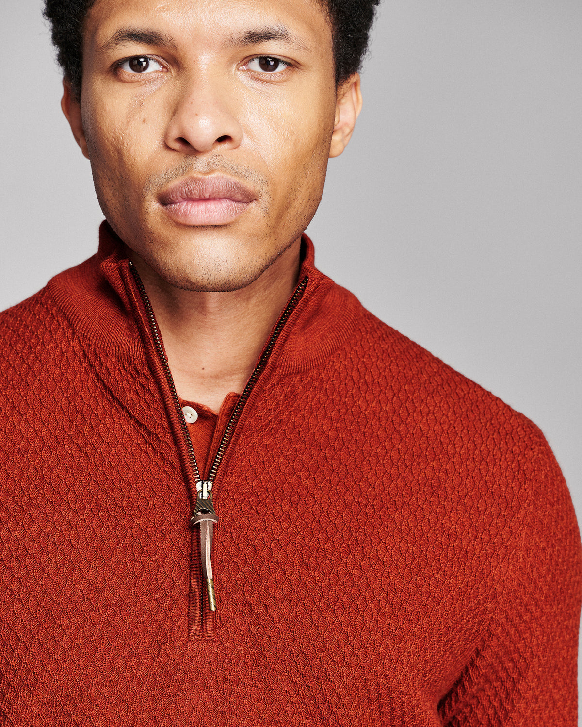 A close-up portrait of a man wearing a textured rust-orange quarter-zip knit sweater against a solid gray studio background.