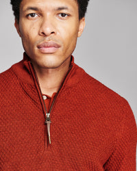 A close-up portrait of a man wearing a textured rust-orange quarter-zip knit sweater against a solid gray studio background.