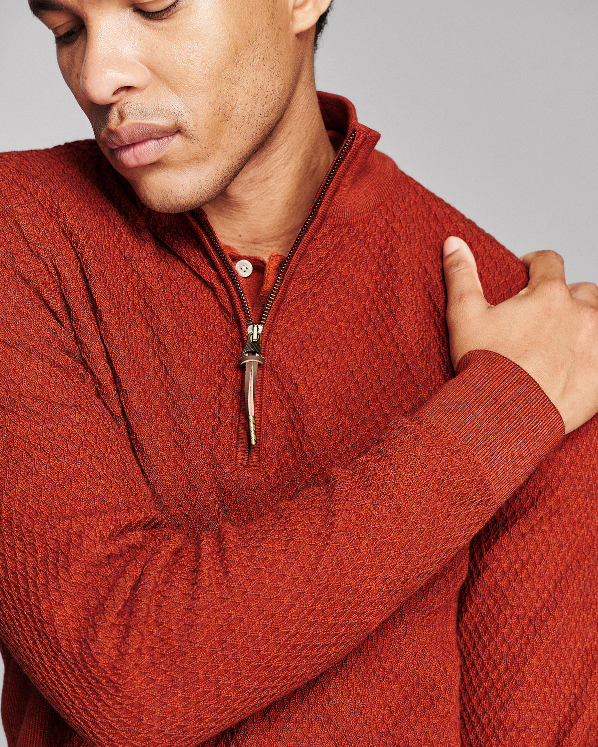 Close-up of a man wearing a textured rust-orange quarter-zip pullover sweater, showing the ribbed collar, zipper, and long sleeves against a neutral gray background.