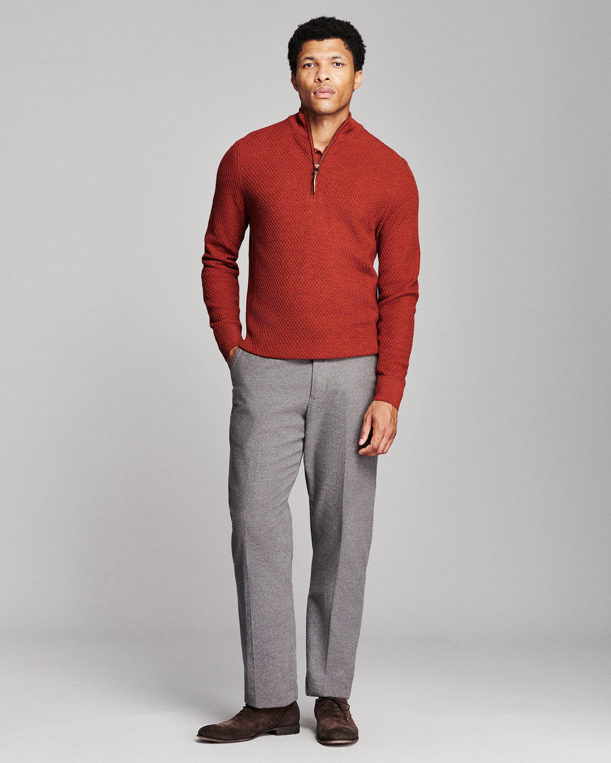 A man modeling a textured rust-red quarter-zip sweater and gray trousers against a light gray studio background.