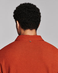 Back view of a person with curly black hair wearing a textured rust-orange polo shirt against a light gray studio background.