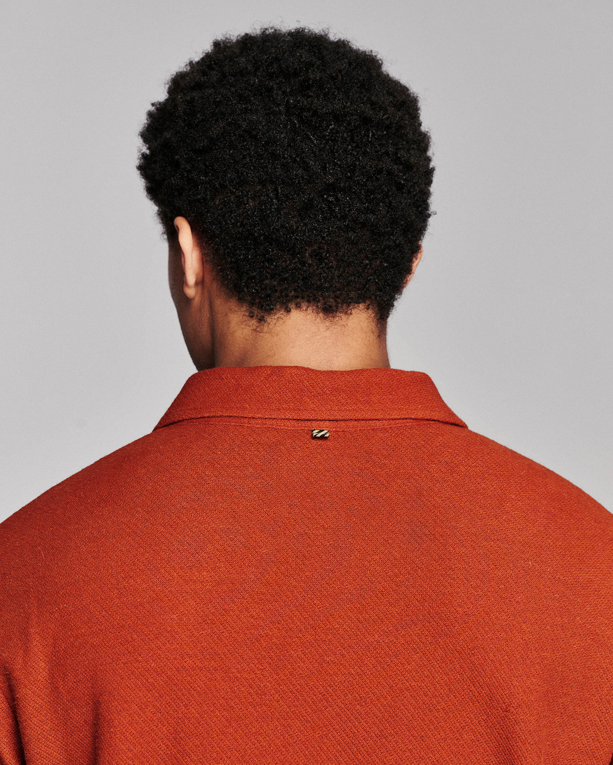 Back view of a person with curly black hair wearing a textured rust-orange polo shirt against a light gray studio background.